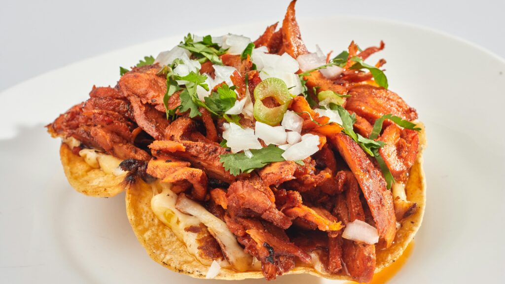 Close-up of a flavorful tostada topped with marinated meat, fresh onions, and herbs on a white plate.